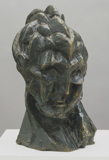 A dark green, bronze-like sculpture of a distorted human face features sharp, angular shapes and uneven surfaces. The piece is textured with gold accents and displayed on a white platform. This is an AI generated description. To give feedback, email ai@moma.org.