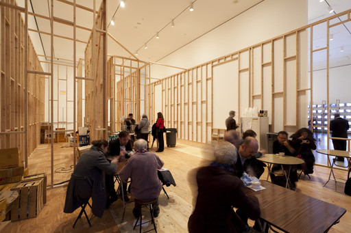 A spacious room with exposed wooden framing and light-colored walls, filled with several groups of people sitting at small tables and engaged in conversation. The lighting is bright, and the floor is made of wood. This is an AI generated description. To give feedback, email ai@moma.org.