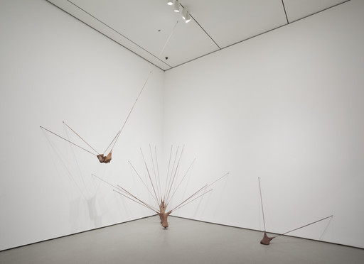 A metallic sculpture with long, slender rods extends dynamically from the floor and wall. Predominantly brown tones contrast with the plain white background. This is an AI generated description. To give feedback, email ai@moma.org.