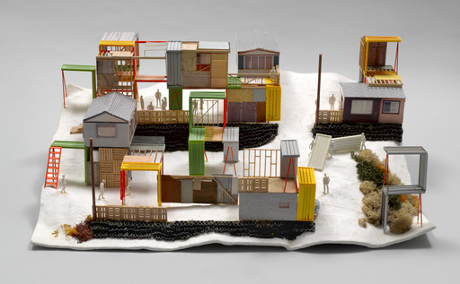 The image shows a model of a miniature village with structures made of multicolored materials, including gray, yellow, green, and red elements, on a white surface. Small figurines are positioned around the structures. This is an AI generated description. To give feedback, email ai@moma.org.