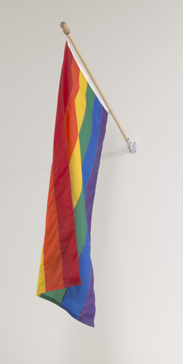 A colorful flag with horizontal stripes of red, orange, yellow, green, blue, and purple hangs from a wooden pole attached to a wall. The background is plain and light-colored. This is an AI generated description. To give feedback, email ai@moma.org.