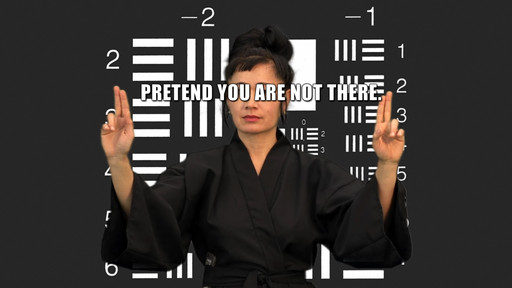 A person in a black robe holds up both hands in front of a dark background with white geometric patterns and the text "PRETEND YOU ARE NOT THERE." The image predominantly features black and white colors. This is an AI generated description. To give feedback, email ai@moma.org.