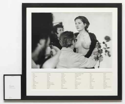 A black-and-white photo shows a person standing topless while others interact closely around them. Below the photo, there is a list of items including "gun," "rose," and "wine" in black text on a white background. This is an AI generated description. To give feedback, email ai@moma.org.