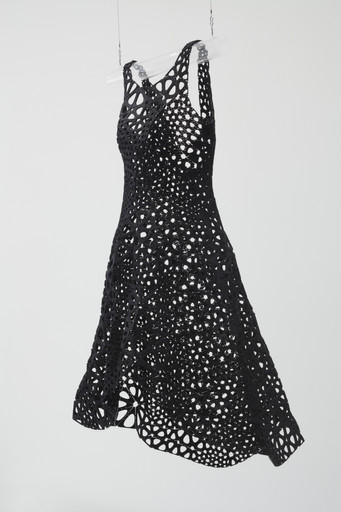 A black dress with numerous circular and irregular cutouts hangs suspended on a clear hanger against a plain white background. The dress flares out slightly at the bottom. This is an AI generated description. To give feedback, email ai@moma.org.