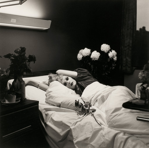 A person lies in a bed with white linens, surrounded by flowers in vases on either side. The room is dimly lit with a light fixture above, and the person has an arm resting on their head. This is an AI generated description. To give feedback, email ai@moma.org.