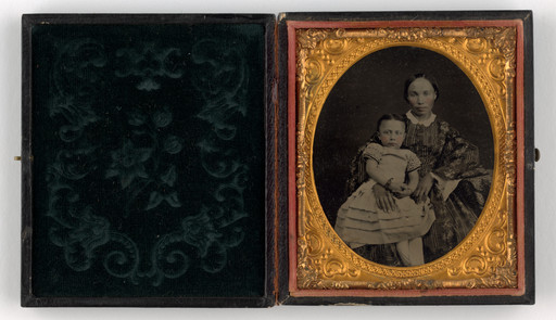 A vintage photograph shows an adult and a child seated together in a gold oval frame. They both wear patterned clothing with predominantly dark shades, in a case with intricate floral designs. This is an AI generated description. To give feedback, email ai@moma.org.