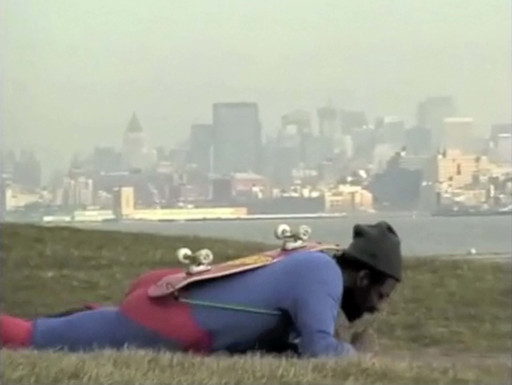 A person dressed in a blue and red outfit lies prone on a grassy area with a skateboard strapped to their back. The background features a hazy city skyline with several high-rise buildings. This is an AI generated description. To give feedback, email ai@moma.org.
