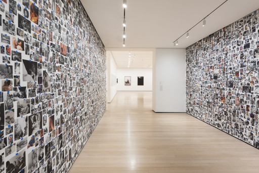A hallway with light wood floors is lined with hundreds of black-and-white and colored photos covering both walls. The ceiling has rows of spotlights, and the hallway opens into a white-walled room with framed artworks. This is an AI generated description. To give feedback, email ai@moma.org.
