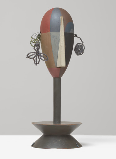 A sculpture features a head with geometric patterns in red, blue, brown, gray, and white, mounted on a dark pedestal. Coiled wires extend from the sides, resembling ears or decorations. This is an AI generated description. To give feedback, email ai@moma.org.