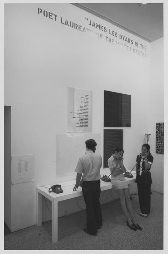 Three people use rotary phones at a white table against a white wall with black and white text posters. The wall also has a sign stating, "James Lee Byars is the Poet Laureate of the United States." This is an AI generated description. To give feedback, email ai@moma.org.