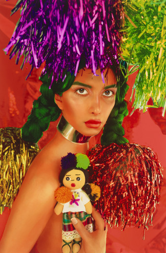 A person with braided green hair holds a small doll, surrounded by colorful tinsel in purple, green, and red. The background is vibrant red, and the person wears a metallic choker. This is an AI generated description. To give feedback, email ai@moma.org.