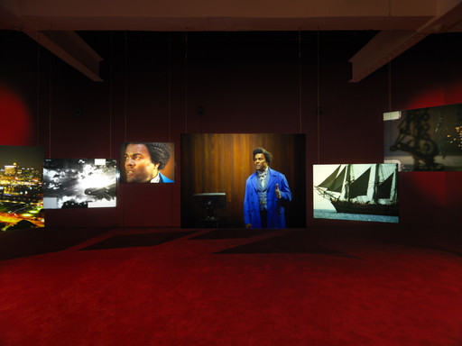 The image shows five photographs hanging on a dark wall, each lit individually. These include city lights, a person in blue clothing, a sailing ship, and a black-and-white scene; the floor is red. This is an AI generated description. To give feedback, email ai@moma.org.